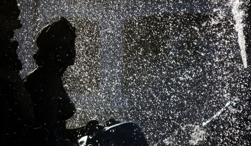 A classical statue silhouetted against glittering, backlit fountain spray