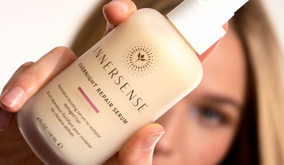 A woman holds a bottle of Innersense Overnight Repair Serum close to the camera, with her face softly blurred in the background.