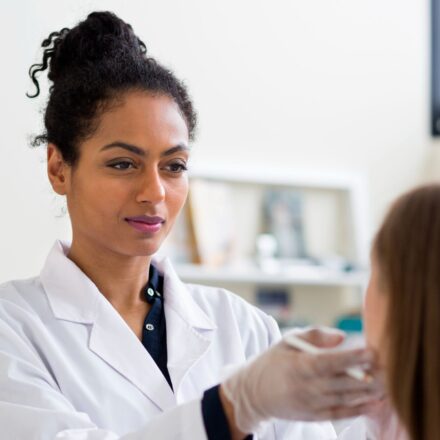 A clinician wearing a white coat and gloves examines a patient’s face during a consultation.
